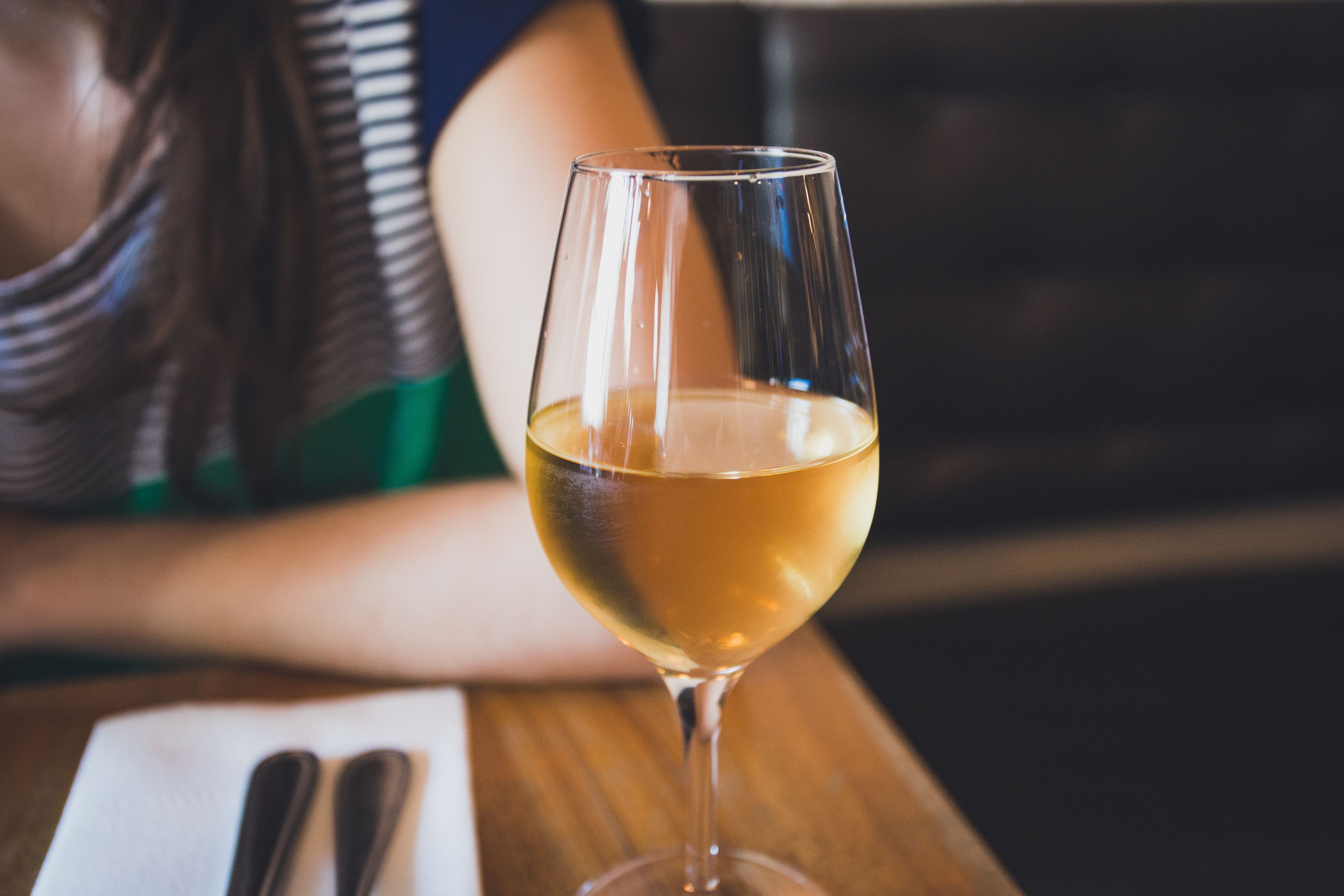 white wine in a glass on a table with arm of woman who is seated at table in background 