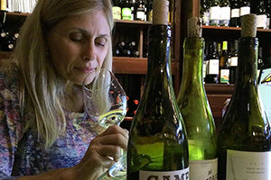 Woman with blond hair and multicolored shirt looking into and sniffing a glass of white wine with three bottles of wine in the foreground and  shelves of wine in the background